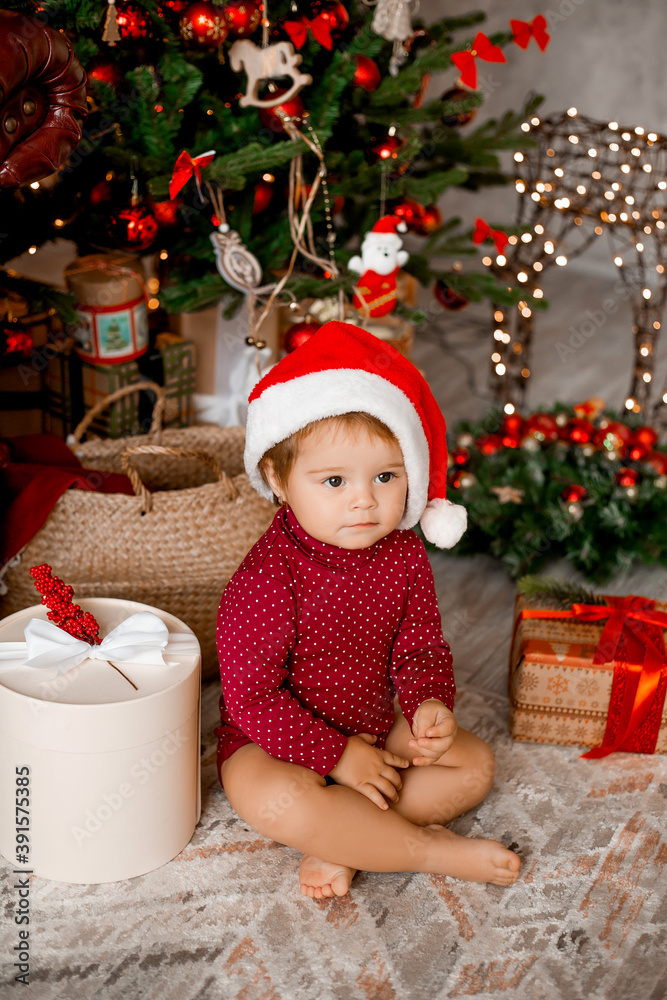Cute baby Santa sits at home near the Christmas tree with gifts Cute baby Santa sits at home near the Christmas tree with gifts