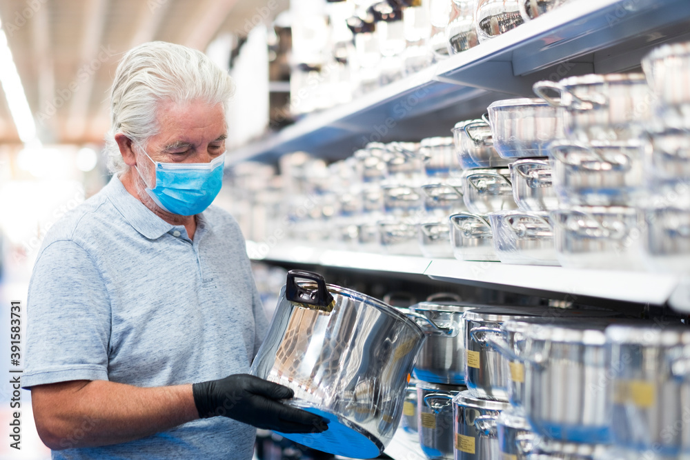 Obraz premium A white haired senior man looking at household steel pans in a store wearing a surgical mask due to the coronavirus covid-19. Consumerism concept