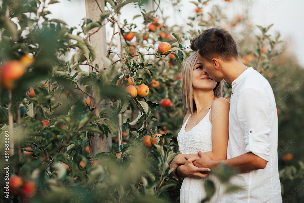 Happy young couple in white clothes in apple orchard. They looking each other and man hugging his girl from back.