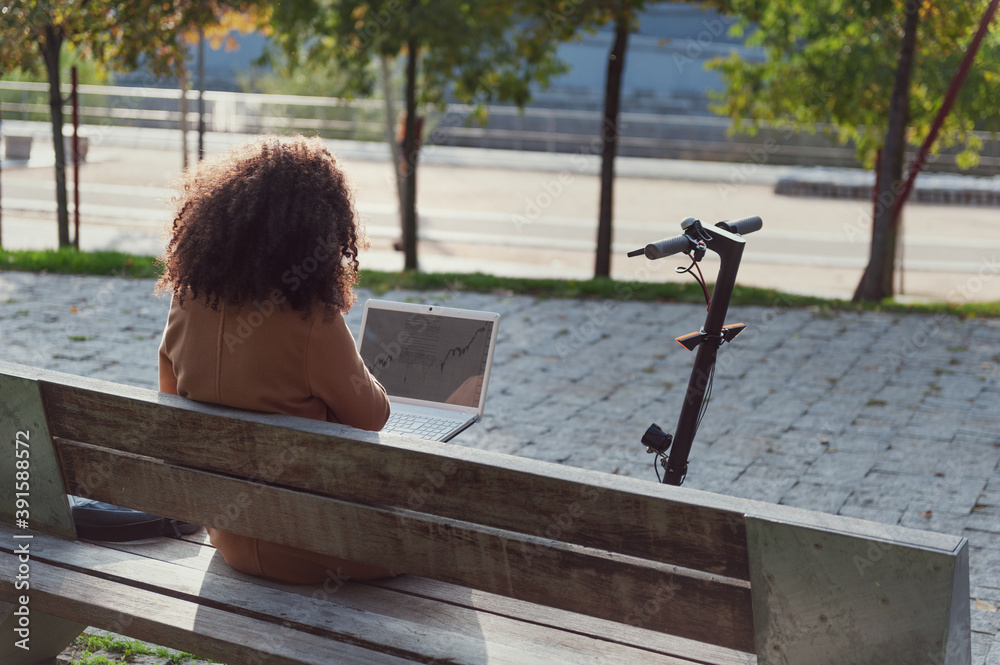 young woman sitting on a bench with a laptop and an electric scooter