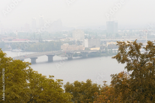 Kyiv, Ukraine-November 03, 2020:Autumn foggy morning landscape view of Dnipro River with The Harbour Bridge. Building in morning mist on the horizon. Tree Leaves Border. Natural Frame
