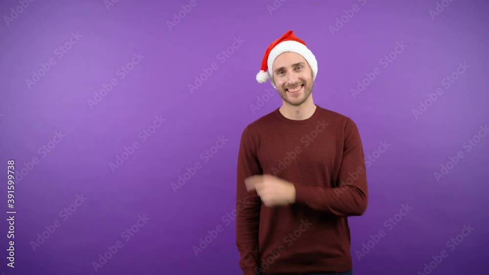 young attractive bearded man in a christmas santa hat points his finger next to the blank space. man over isolated purple background. 4K young attractive bearded man in a christmas santa hat points his finger next to the blank space. man over isolated purple background. 4K