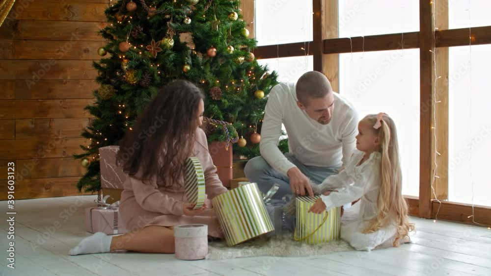 Happy family looking gifts over Christmas tree on holiday morning. Mod, dad and two daughter on floor opening gift boxes on decorated New Year tree background in living room