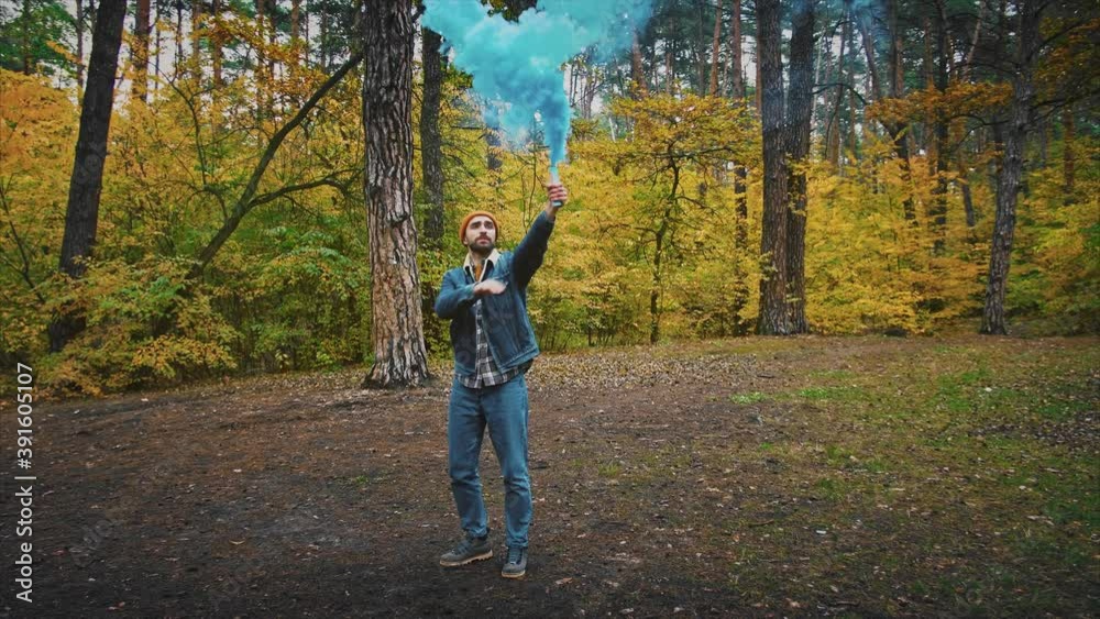 Young man in forest having fun with blue smoke grenade in autumn forest, 360 degree tracking arc shot