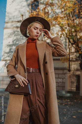 Outdoor autumn fashion portrait of elegant, luxury woman wearing trendy midi beige, camel color coat, brown hat, orange turtleneck, with leather trapeze shoulder bag, walking in street of city