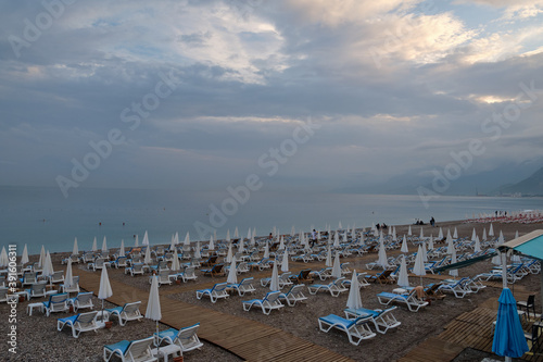 Deserted beach in Antalya. Sea, beautiful sky. Lots of white sunbeds with blue pillows, folded umbrellas. In the distance, figures of people. End of the season. © Aleksandr