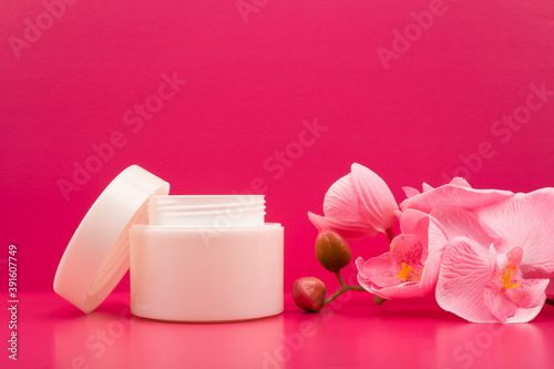 Cosmetic cream in a white jar and flowers on bright pink background with space for text