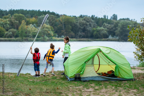 Three children standing on river bank near tent in camping site and playing