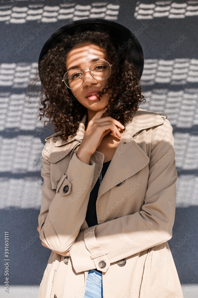 Stylish black girl with curly hair glasses black hat and beige raincoat. American black lady street style casual modern city walk industrial look Stock Foto Adobe Stock