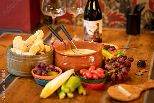 A pan of cheese fondue on a table with fresh vegetables, fruits and a bottle of wine