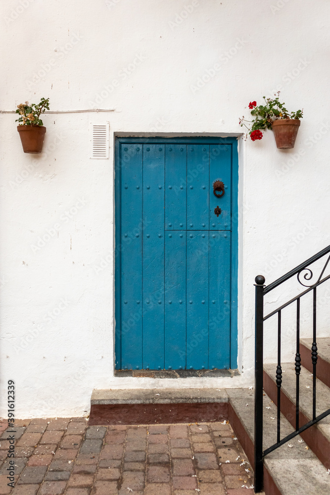 Fototapeta premium Blue wooden door on a white facade, typical house in a Spanish traditional village. Closed door with a metal handle, flower pots with geraniums hanging from the wall. Almería, Andalusia, South Spain