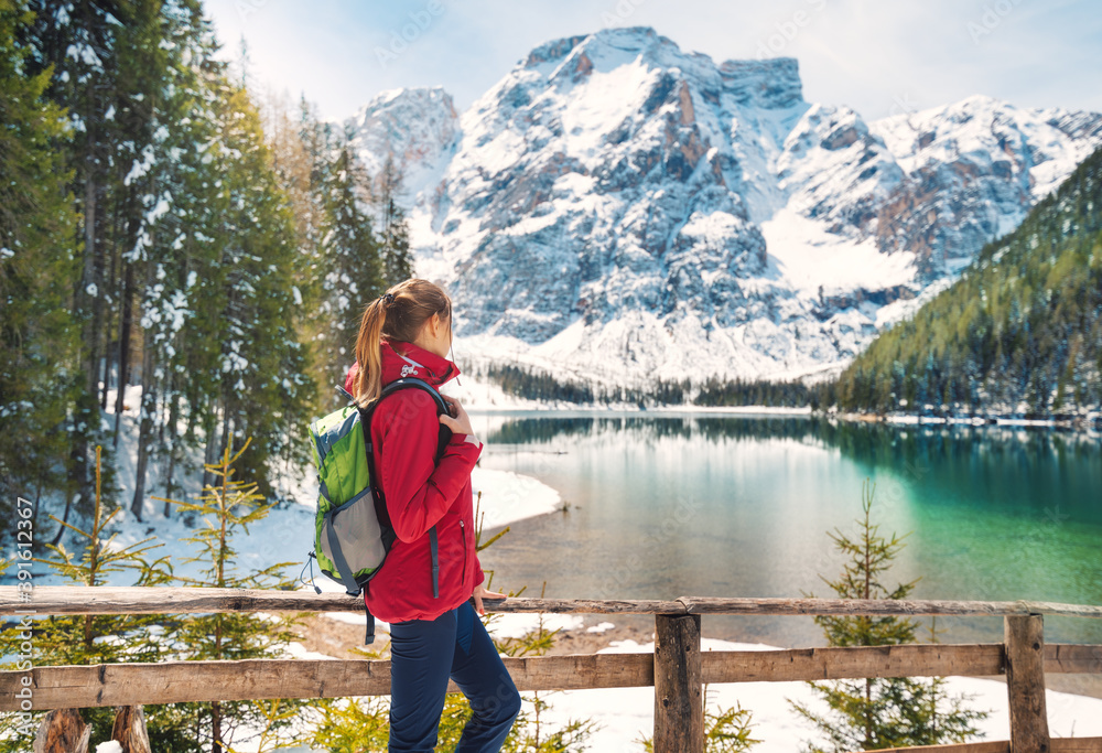 Young woman with backpack near wooden fence and snowy shore of Braies lake with clear water at sunny day in winter. Travel. Landscape with slim girl, reflection in water, mountains in snow, trees