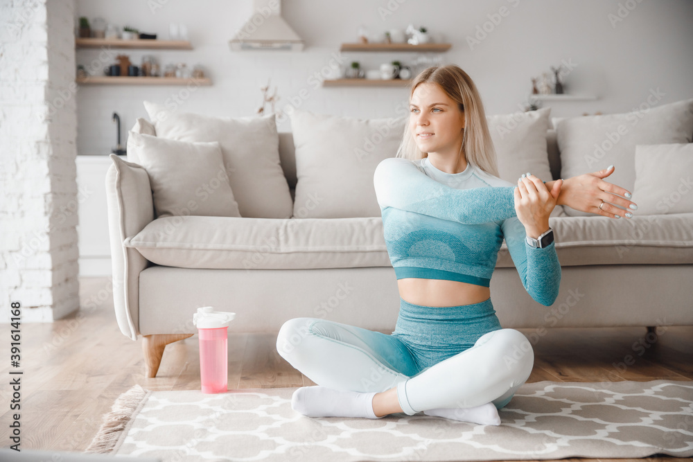 Young woman stretching indoors at home with smart watch. Protein shake or bottle of water stay around sofa
