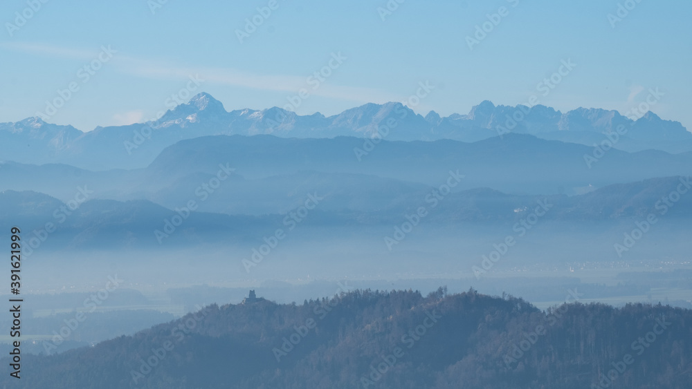 mystical scenery with layers of mountains in a beautiful landscape with hilltops softly peaking out of white clouds. Castle ruins on hilltops and the Julian alps with mount Triglav in the background. 