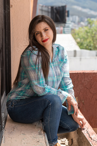 white skinned latin person with long hair and red lips, happy and sitting , wearing a plaid t-shirt and jeans