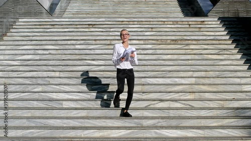 A girl in a white shirt and with a pack of papers in her hands goes down the street stairs, jumping up, and throws the sheets of paper up in a dance. Happy business woman. Slow motion at 120 fps. photo