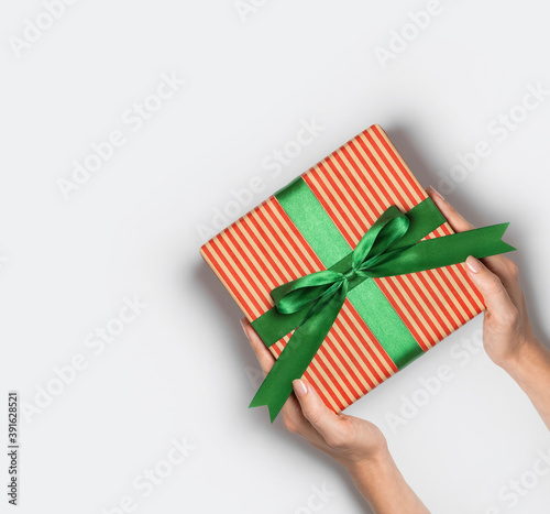 Female hands hold gift box in striped craft paper with green bow on light background flat lay top view copy space. Holiday present, christmas, new year, birthday, valentine's day, 8 march. Surprise