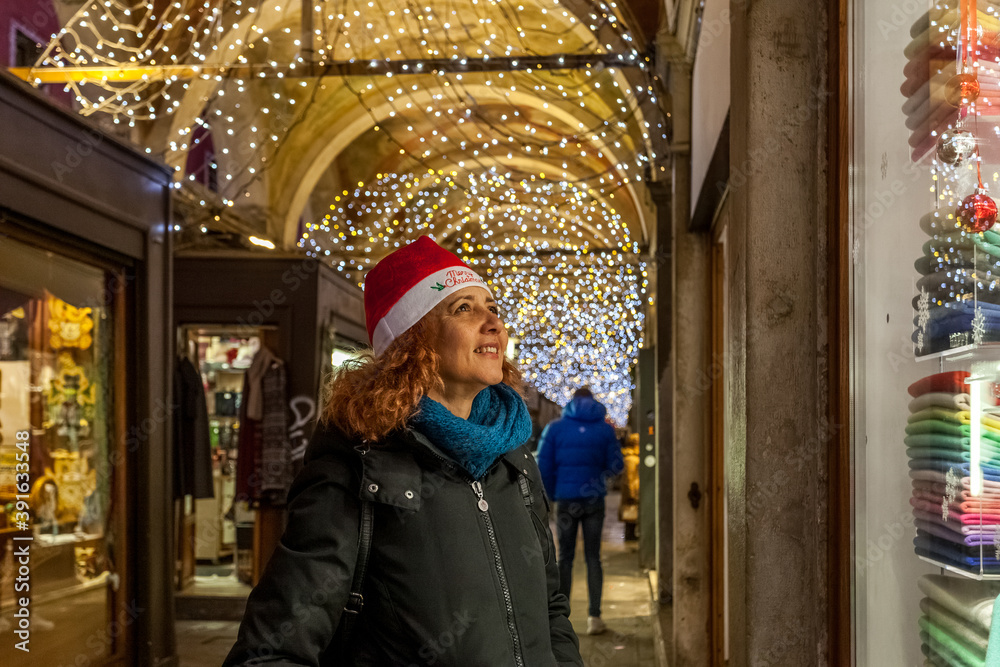 Brazilian woman with Christmas hat looks smiling at an illuminated shop window in a street of Venice, Italy