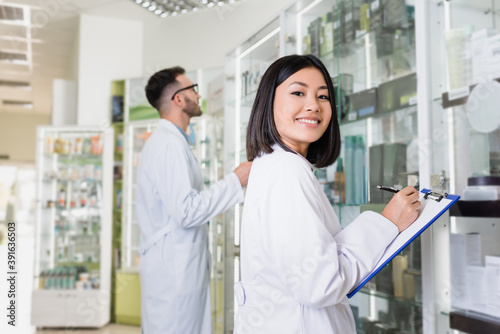happy asian pharmacist in white coat writing on clipboard near colleague on blurred background