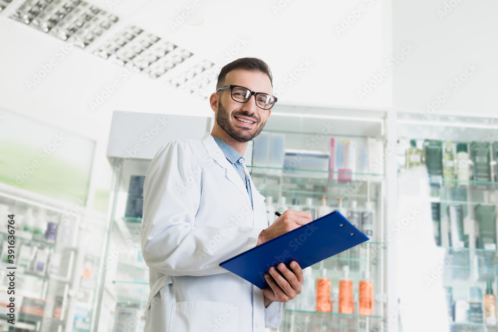cheerful pharmacist in eyeglasses and white coat writing on clipboard in drugstore