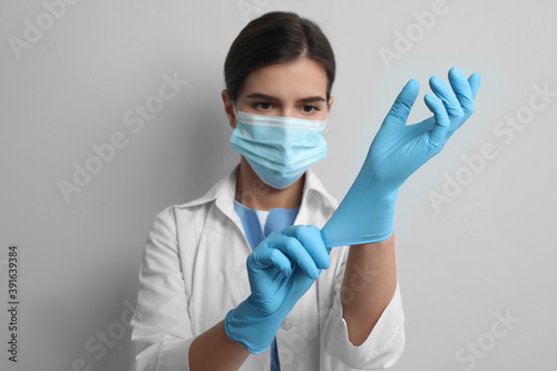Doctor in protective mask putting on medical gloves against light grey background