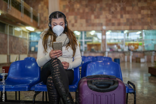 Young woman sitting on train station with face mask and luggage while listening music