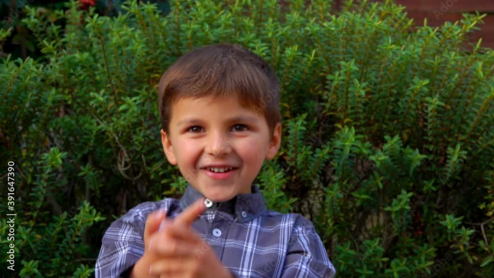 The cheerful little boy in a plaid shirt is counting on fingers outdoors on the background of a green bush The cheerful little boy in a plaid shirt is counting on fingers outdoors on the background of a green bush