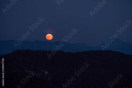 huge orange full moon rising behind a mountain ridge with layers of hills in the foreground creating a mystical atmosphere. glowing super moon rising into the night sky
