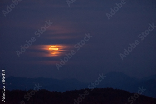 huge orange full moon rising behind a mountain ridge illuminating misty clouds creating a mystical atmosphere. glowing super moon rising into the night sky with layers of hills in the foreground.
