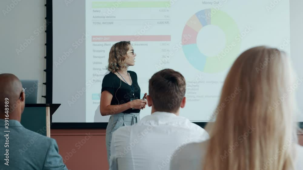 Business woman pointing at projection screen and explaining the statistics to the audience. Female speaker giving an informative presentation at conference.
