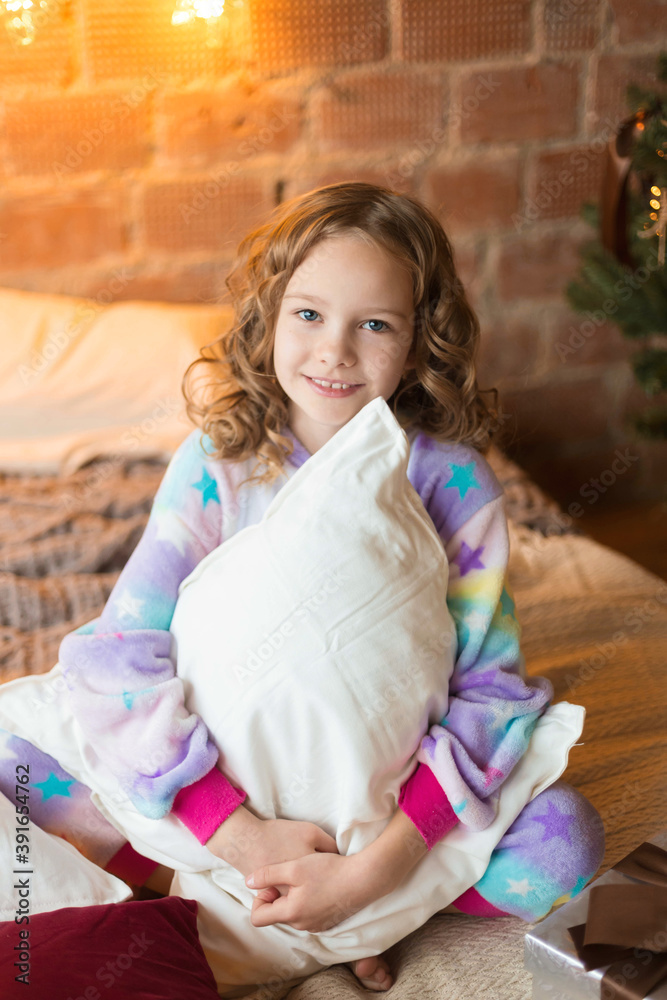 Fototapeta premium Cute little girl sits on the bed and huggs pillows with background of tree