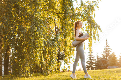 Portrait cheerful young yoga woman holding yoga mat at city park, looking away. Active young woman holding yoga mat and smile at park, ready to practice yoga with happiness feeling