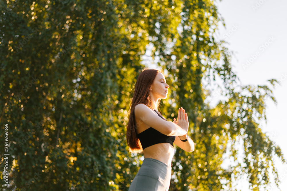 Low angle shot of meditative redhead woman practicing yoga performing making hand greeting namaste in city park background of bright sunray and green grass in morning at dawn. Low angle shot of meditative redhead woman practicing yoga performing making hand greeting namaste in city park background of bright sunray and green grass in morning at dawn.