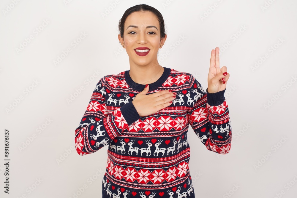 Young beautiful Arab woman wearing Christmas sweater, against white background smiling swearing with hand on chest and fingers up, making a loyalty promise oath. Young beautiful Arab woman wearing Christmas sweater, against white background smiling swearing with hand on chest and fingers up, making a loyalty promise oath.