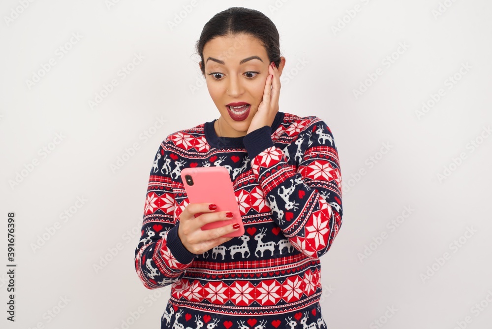 Young beautiful Arab woman wearing Christmas sweater against white wall, holding in hands cell reading sms using new app 5g Young beautiful Arab woman wearing Christmas sweater against white wall, holding in hands cell reading sms using new app 5g