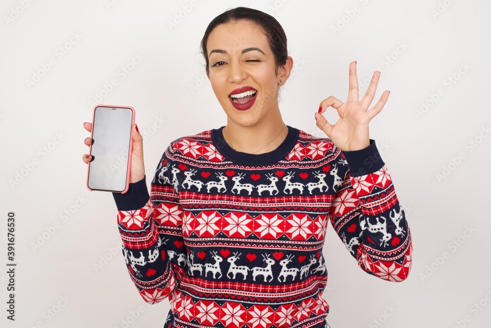 Young beautiful Arab woman wearing Christmas sweater against white wall, holding in hands cell showing ok-sign Young beautiful Arab woman wearing Christmas sweater against white wall, holding in hands cell showing ok-sign