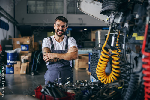 Smiling happy bearded tattooed worker in overalls standing next to truck with arms crossed.