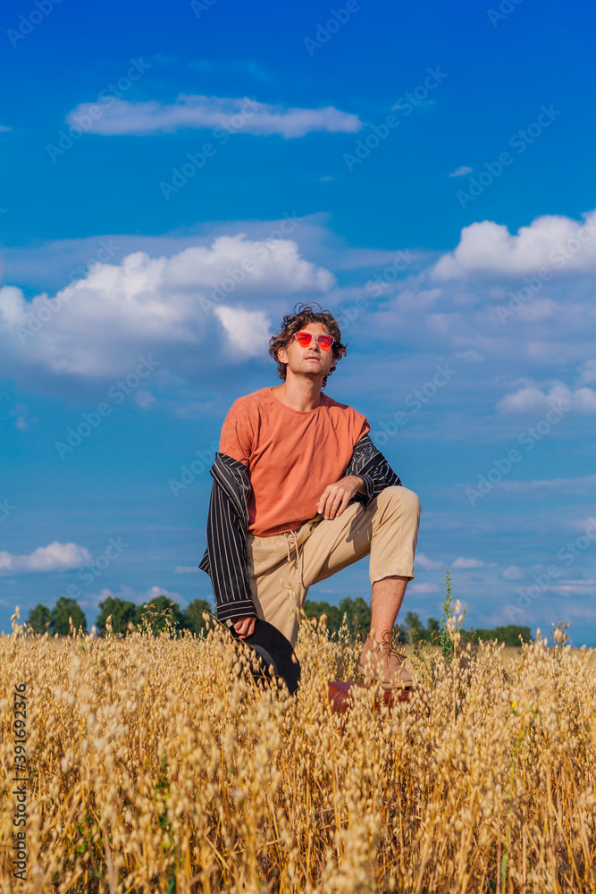 Tall handsome man with black hat and pink sunglasses standing with one leg on a brown vintage leather suitcase at golden oat field. Tall handsome man with black hat and pink sunglasses standing with one leg on a brown vintage leather suitcase at golden oat field.