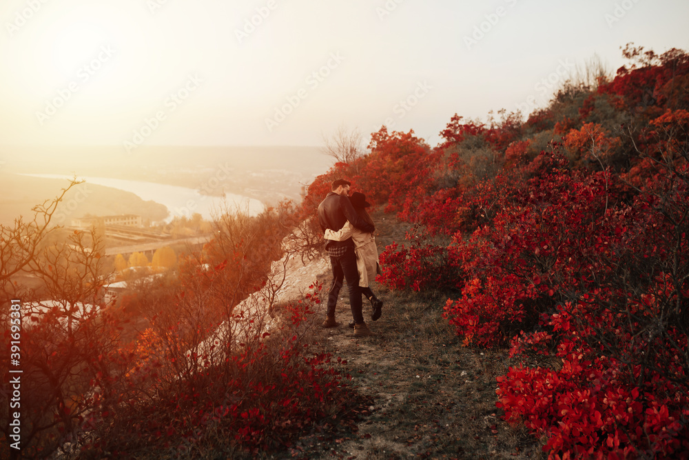 Romantic couple standing and hugging on a autumn hill with beautiful landscape. They watching the sunset. Back view.