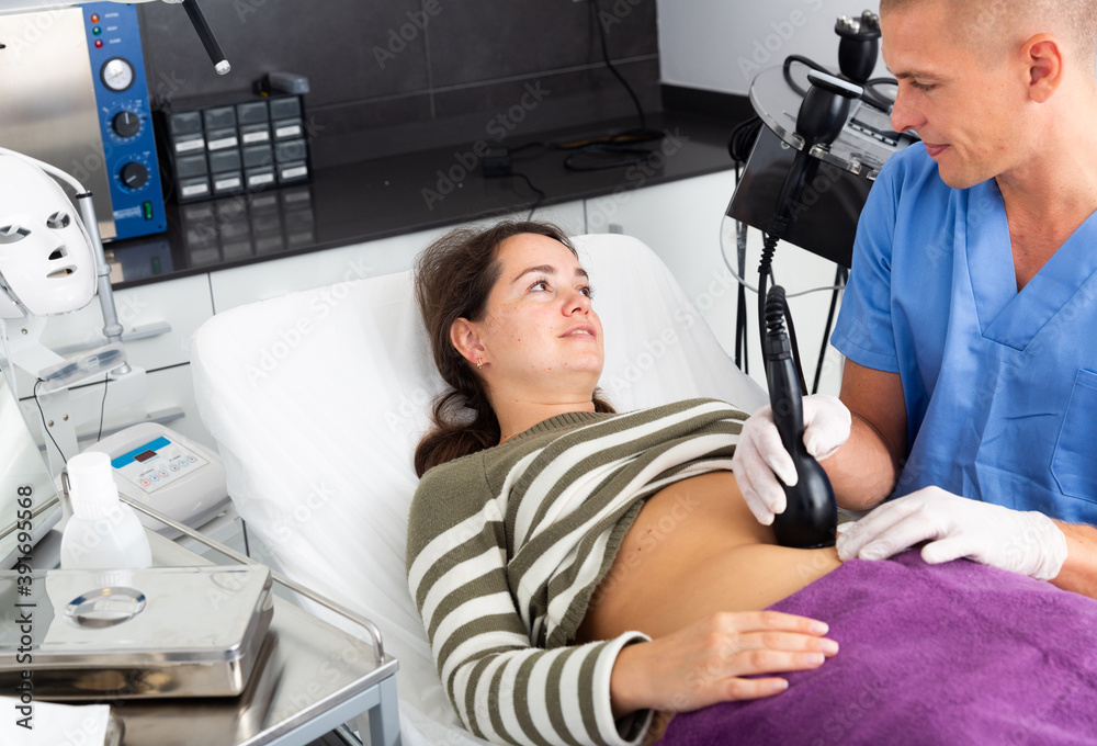Obraz premium Confident male doctor performing radiofrequency lifting procedure on abdomen of young woman, new technologies for slim lifting body
