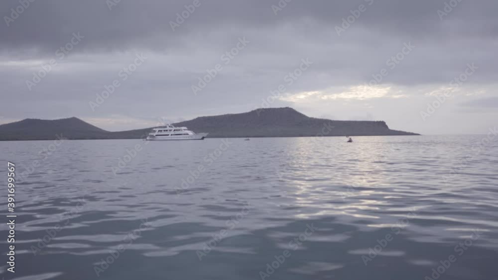 A kayak with two tourists approaches a cruise that is anchored near an island of the Galapagos islands. The ocean is reflecting the sunlight. A kayak with two tourists approaches a cruise that is anchored near an island of the Galapagos islands. The ocean is reflecting the sunlight.