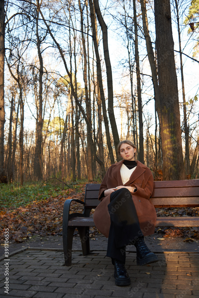 Fototapeta premium A charming girl with blonde hair , wearing a brown wool coat, is sitting on a wooden bench in an alley in an autumn Park.