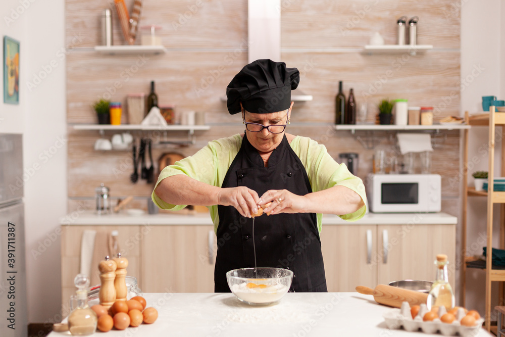 Elderly woman preparing dough cracking eggs over wheat flour following traditional recipe. Elderly pastry chef cracking egg on glass bowl for cake recipe in kitchen, mixing by hand, kneading. Elderly woman preparing dough cracking eggs over wheat flour following traditional recipe. Elderly pastry chef cracking egg on glass bowl for cake recipe in kitchen, mixing by hand, kneading.