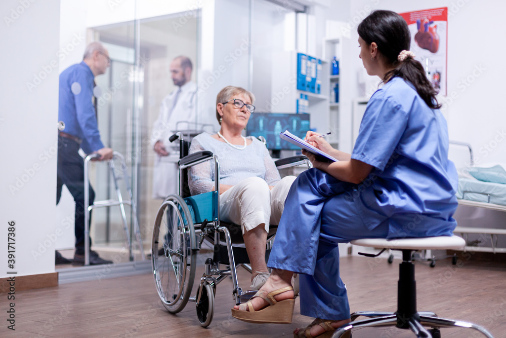 Handicapped woman in wheelchair answering nurse questionnaire during medical examination in hospital room. Help for disabled people. Elderly assistance for old man with walking frame. Handicapped woman in wheelchair answering nurse questionnaire during medical examination in hospital room. Help for disabled people. Elderly assistance for old man with walking frame.