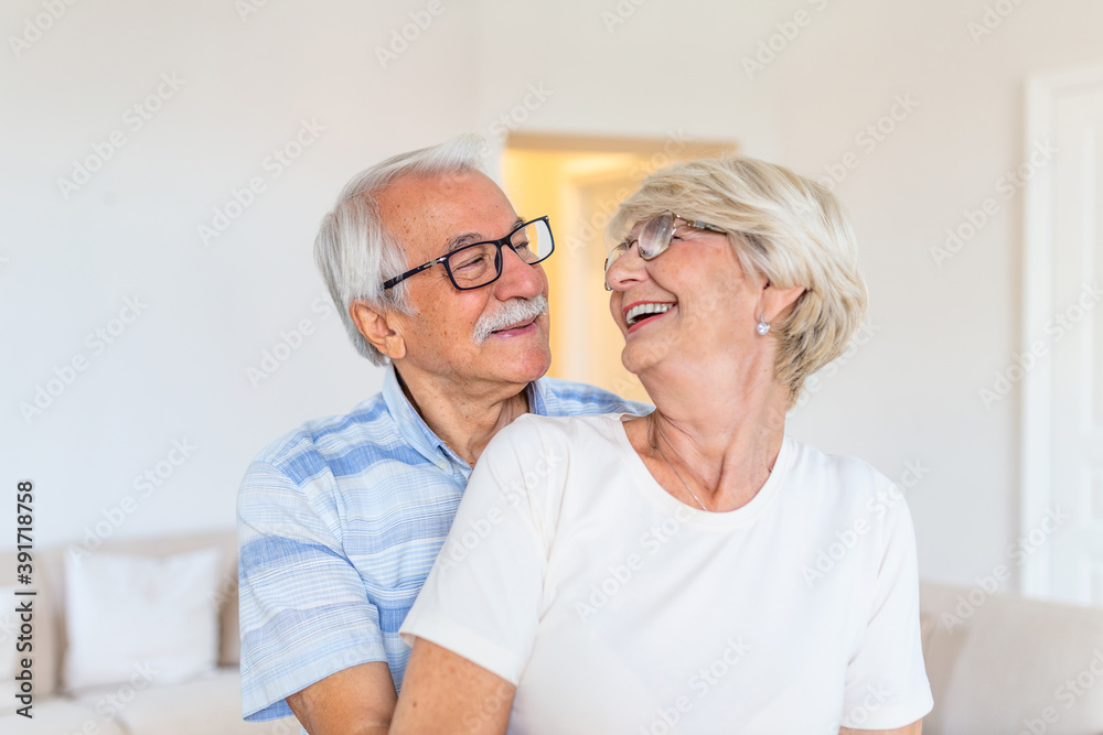 Elderly couple standing in the living room. Portrait of happy senior couple embracing each other in living room at home. Old couple looking each other with love and smilling. Elderly couple standing in the living room. Portrait of happy senior couple embracing each other in living room at home. Old couple looking each other with love and smilling.