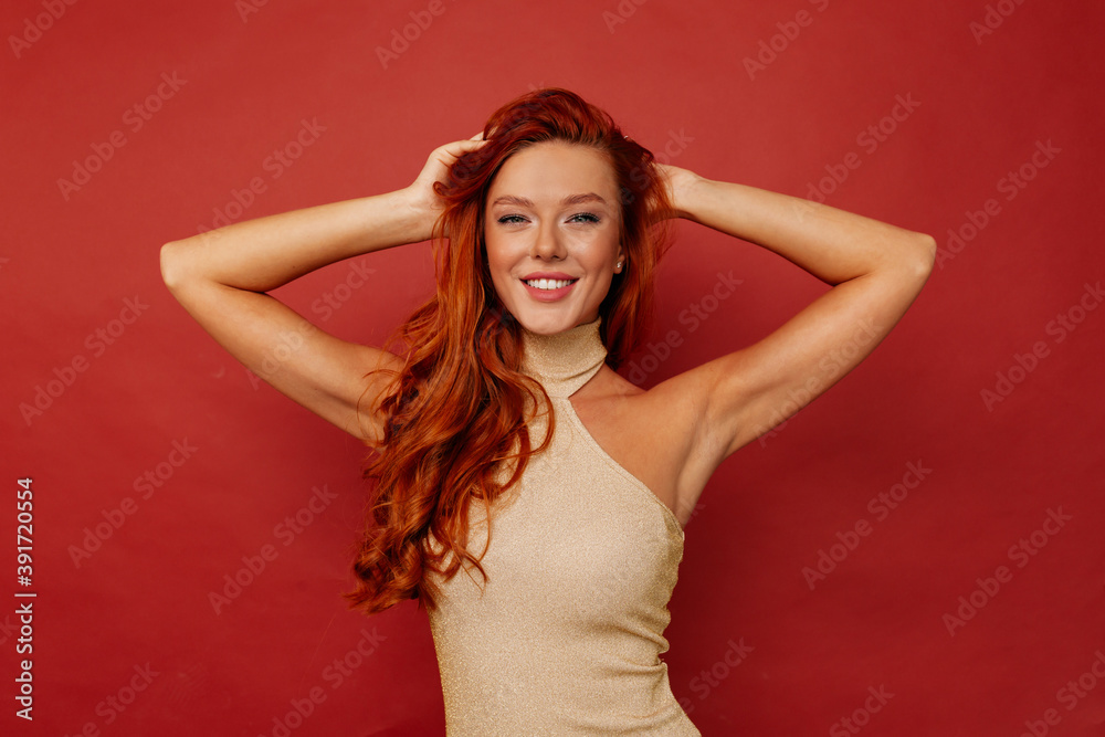 Sensual woman with long red hair touching her hair and smiling at camera on isolated background. Studio shot of glamorous lady in dress isolated on red background. Sensual woman with long red hair touching her hair and smiling at camera on isolated background. Studio shot of glamorous lady in dress isolated on red background.