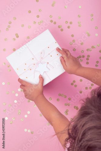 Top view of child hands holding pink gift box with bow and star over  background decorated with golden confetti. Flat lay holiday festive concept. Authentic real life moments. photo