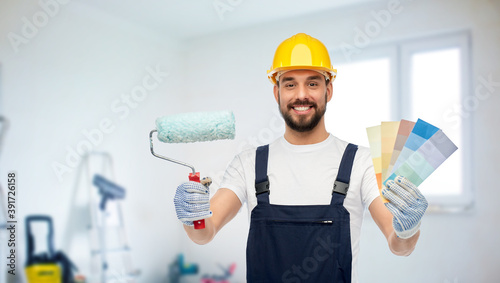 repair, construction and building concept - happy smiling male worker or builder in yellow helmet and overall with paint roller and color palettes over room with equipment on background photo