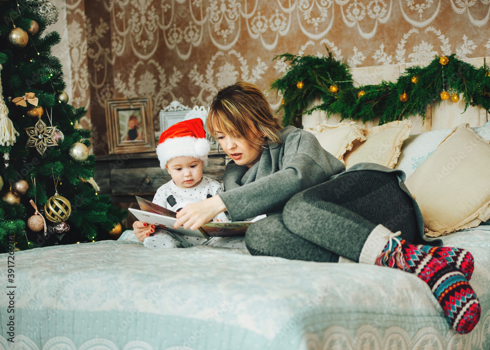 Merry Christmas and Happy Holidays. Cheerful mom and her cute son reading the book. Parent and little child having fun near Christmas tree indoors. Morning Xmas. Portrait family close up. Merry Christmas and Happy Holidays. Cheerful mom and her cute son reading the book. Parent and little child having fun near Christmas tree indoors. Morning Xmas. Portrait family close up.