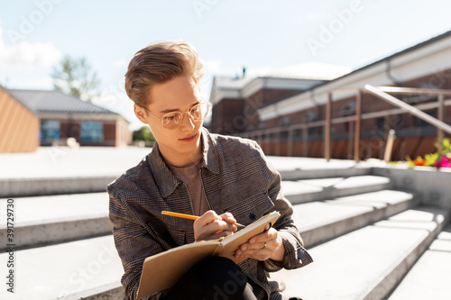 leisure, education and people concept - young man or teenage boy in glasses with notebook, diary or sketchbook writing or drawing in city